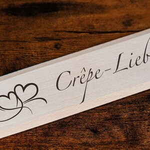Könnte beinhalten: Ein Holz-Crêpe-Spatel mit dem Text "Crêpe-Liebe" und einem stilisierten Herz-Design.