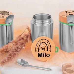 Könnte beinhalten: Isolierte Lebensmittelbehälter aus Edelstahl mit Holzdeckeln. Ein Deckel zeigt ein Igel-Design und den Namen "Lia", ein anderer ein Bär-Design, und der dritte ein Regenbogen-Design mit dem Namen "Milo". Ein kleiner Löffel ist enthalten.