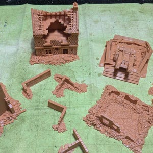 Op de afbeelding: Een verzameling ongeverfde, lichtbruine miniatuur terreinstukken voor tabletop-spellen, waaronder een verwoeste toren, een beschadigd huis, diverse muursegmenten en met puin gevulde gebouwfunderingen met gedetailleerde baksteen- en steentexturen.