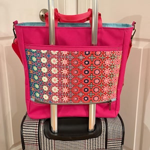 May include: A pink tote bag with a colorful patterned pocket and a zippered closure. The bag is attached to a black rolling suitcase with a black and white checkered pattern.