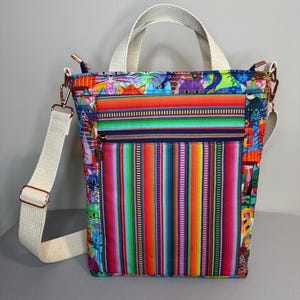 May include: A colorful tote bag with a vibrant striped pattern in shades of red, green, yellow, and blue. The bag features a front zippered pocket, a cream-colored shoulder strap, and matching handles. The bag's sides have a whimsical cat print.