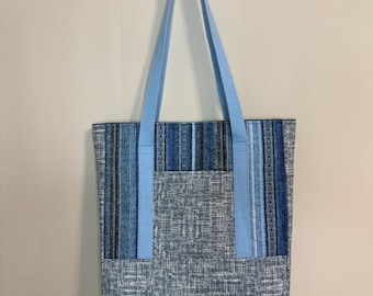 May include: A blue tote bag with light blue straps hanging from a white hook. The bag features a textured, denim-like fabric with vertical stripes of varying blue shades. A light blue pocket is on the front.