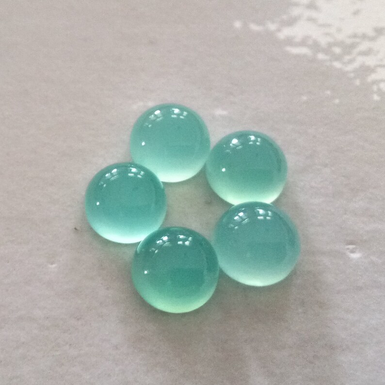 May include: Six round, light blue cabochon gemstones on a white surface.
