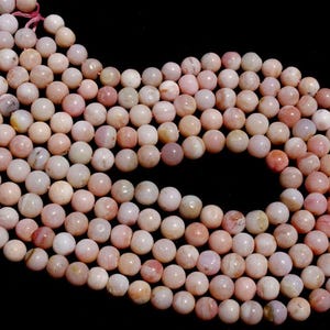 May include: Strands of round, light pink gemstone beads. The beads have a smooth, polished surface and vary slightly in color, with some showing darker pink and peach tones. The beads are strung together with pink thread, and the background is black.