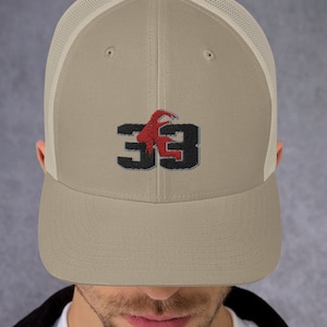 Könnte beinhalten: Beige Trucker-Cap mit Mesh-Rückseite und gebogenem Schirm. Auf der Kappe ist die Zahl "33" in Schwarz mit einer roten Grafik abgebildet. Die Kappe wird von einer Person getragen.