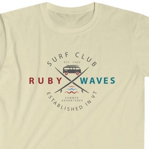 Könnte beinhalten: Hellbeige T-Shirt mit einem kreisförmigen Aufdruck. Es zeigt "Surf Club Ruby Waves" mit einem Vintage-Van, gekreuzten Surfbrettern und Wellen. Der Text "Established in VT" ist ebenfalls enthalten.