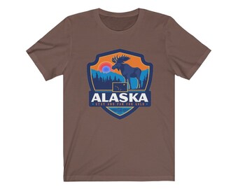 Puede incluir: Una camiseta marrón con un gráfico azul y naranja de un alce frente a una cordillera. El gráfico también incluye una puesta de sol y el texto "Alaska - Stay and Pan for Gold - ".
