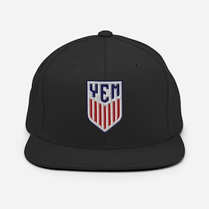 May include: A black baseball cap featuring an embroidered shield-shaped patch on the front. The patch displays the letters YEM in blue at the top, positioned above a series of vertical red and white stripes, all outlined in a dark blue border.