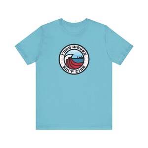 Könnte beinhalten: Ein hellblaues T-Shirt mit einem runden Logo, das eine rote Welle, einen Leuchtturm und den Text "Ruby Waves Surf Club" zeigt.