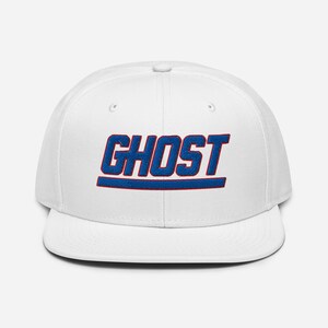 May include: A white snapback cap featuring the word GHOST embroidered in bold blue letters with a red outline. The text is centered on the front panel, accompanied by a horizontal blue line with a red outline positioned directly underneath.