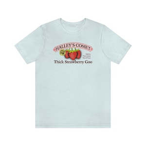 May include: A light blue t-shirt with a pink and black graphic of a label that reads "Halley's Comet Thick Strawberry Goo" with a cartoon strawberry and a logo that says "Good Goo" in a circle.