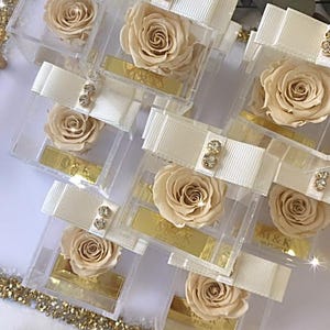 Op de afbeelding: Verschillende transparante acrylboxen, elk met een geconserveerde crèmekleurige roos. De dozen zijn versierd met witte strikken, versierd met kleine, sprankelende strass-steentjes. Een goudkleurig rechthoekig label is zichtbaar in elke doos.