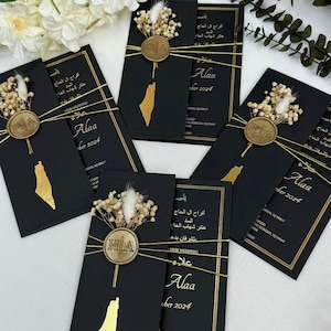 May include: A collection of black wedding invitations with gold detailing. Each card has a gold map outline, dried flowers, and a gold seal with the initials "HA". The invitations are tied with gold string and include Arabic text.