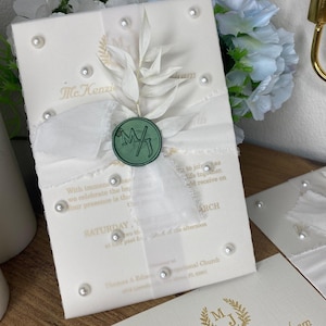 May include: Ivory wedding invitation with gold text, a white ribbon, and a green wax seal. The invitation is embellished with faux pearls and a sprig of dried foliage. The text reads "McKenzie Eden Graham" and "Saturday, March".