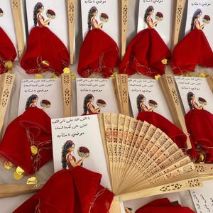 May include: A display of decorative fans and cards. The fans are crafted from light brown wood, featuring detailed cut-out patterns. Each card showcases an illustration of a woman in a red dress. The text on the cards is in Arabic.