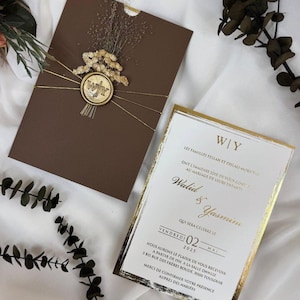 May include: A brown wedding invitation with gold detailing. The invitation features the initials "W|Y" and the names "Walid & Yasmine" in gold script. The invitation is decorated with dried flowers and gold string.