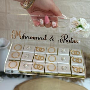 Puede incluir: Una caja de acrílico transparente llena de bombones envueltos individualmente. Los bombones tienen elementos decorativos dorados o plateados. La caja tiene los nombres "Muhammad & Rabia" en escritura dorada. Una pequeña flor blanca y un asa transparente están adjuntas.