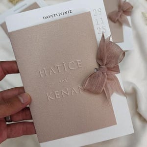May include: A wedding invitation featuring the names "HATICE" and "KENAN" embossed on a textured, light brown panel. The invitation includes the date "29 11 25" and is decorated with a matching ribbon. The word "DAVETLISINIZ" is printed at the top.
