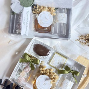 May include: Two gift boxes with clear containers filled with treats, honey, and small bottles. Each box is decorated with ribbons, dried flowers, and labels with initials. The boxes are on a white surface.