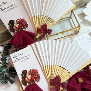 May include: White paper fans with wooden handles and burgundy bows, alongside cards with a woman holding flowers. The cards display the text "Melina Hatice'nin Kına Hatırası". These are decorative items for a special occasion.