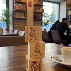 Puede incluir: Tres cubos de madera apilados sobre una mesa de madera. Cada cubo tiene una ilustración de cachimba y el texto "Kuss Ko". El cubo inferior presenta un código QR. Una taza de café está sobre un platillo cerca.