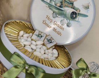 May include: A round, white gift box with a green ribbon and bow, filled with gold-wrapped chocolates and white candies. The lid features a cartoon bear in a pilot uniform on an airplane, with the text "Hoş Geldin Ömer Melih".