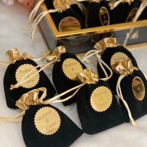 May include: Black velvet pouches with gold drawstring fastenings and gold tags. Each tag is inscribed with the names "Mahmut & Ceren". The pouches are arranged in a decorative display, suggesting a gift or favour.