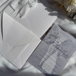 May include: A set of white wedding invitations with embossed details and a ribbon. The invitation suite includes a card with a black and white photo and text. The date "25" is embossed on one envelope.