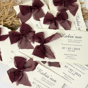 May include: Ivory invitation cards with torn edges, decorated with dark burgundy silk bows. The cards display elegant calligraphy and event details, including a date and time. A stack of cards is arranged on a neutral surface.
