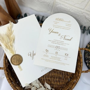 May include: Cream-coloured wedding invitation with gold lettering, featuring the names "Yassin & Nawel" and event details. The invitation is paired with a matching envelope, a wax seal, and dried wheat, presented in a woven basket.