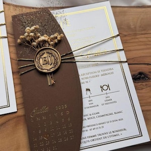May include: Wedding invitation with a brown cover, a calendar for July 2025, and a gold seal with the initials "EJ". The invitation includes details in French, tied with gold string, and decorated with dried flowers.