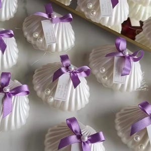 May include: White seashell-shaped favours with purple ribbon bows and pearl accents. Each favour has a small tag attached. The favours are arranged on a gold-trimmed tray, suitable for weddings or special events.