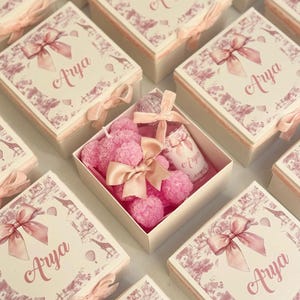 Könnte beinhalten: Eine Sammlung von Geschenkboxen mit rosa Schleifen und dem Namen "Arya" auf den Deckeln. Eine Box ist geöffnet und zeigt rosa rosenförmige Seifen und eine kleine Flasche, ebenfalls mit einer Schleife und dem Namen "Arya". Die Boxen haben ein dekoratives rosa-weißes Muster.