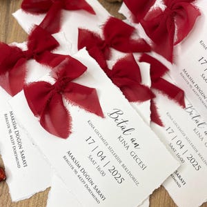 May include: A collection of white invitation cards with torn edges, each topped with a deep red chiffon bow. The cards display elegant calligraphy with event details. The overall design is romantic and celebratory.