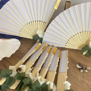 May include: White and beige folding fans with pearl accents and a floral design. Some fans have a gold label with text. The fans are decorated with white calla lilies and green bows. A decorative turtle figurine is also present.