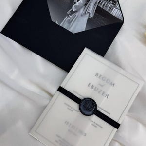 May include: A black envelope containing a black and white photograph of a couple. A translucent invitation card is visible, secured with a black ribbon and a wax seal. The card features text, including names and a date.