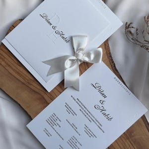 May include: White wedding invitation cards with the names "Dilan & Habib" in elegant script. The cards are tied with a satin ribbon bow. The text "DAVETLISINIZ" is printed at the top. The cards are placed on a wooden surface.