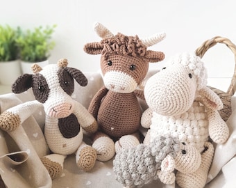 May include: A collection of four crocheted stuffed animals including a black and white cow, a brown highland cow with horns, a large cream-colored sheep, and a small gray sheep, all nestled together inside a woven basket lined with fabric.