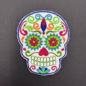 May include: White embroidered sugar skull patch with colorful floral designs, green eyes, pink heart, and green teeth.