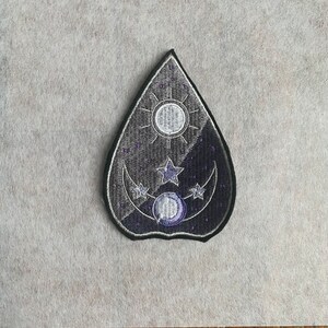 May include: A black and purple embroidered patch in the shape of a teardrop with a crescent moon, stars, and a sun inside.