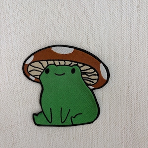 May include: An embroidered patch featuring a bright green frog sitting under a brown mushroom cap with white spots. The frog has a simple black line for a mouth and two black dots for eyes. The mushroom has visible tan gills underneath.