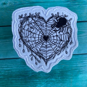 May include: A white heart-shaped patch with black embroidery of a spider web and a spider. The web is in the shape of a heart with a smaller heart in the center.