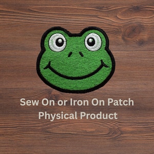 May include: An embroidered patch featuring a green frog face with large white eyes, black pupils, and a wide black smile. The patch has a thick black border. Below the frog, the text reads: Sew On or Iron On Patch Physical Product.