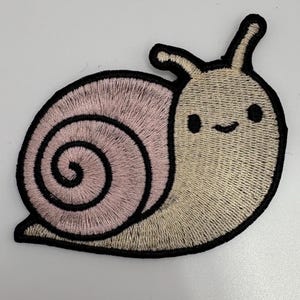 May include: An embroidered patch shaped like a snail with a light pink spiral shell and a beige body. The snail features a black stitched outline, two antennae, and a simple face with two black circular eyes and a small curved line for a mouth.