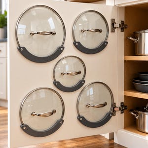 May include: Five circular pot lid organizers mounted on the interior of a cabinet door. Each organizer is a dark gray, crescent-shaped bracket holding a clear glass lid with a metallic handle. The cabinet door is off-white with metal hinges.
