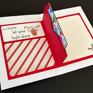 Peut inclure: Une carte de v&oelig;ux de No&euml;l avec un motif &agrave; rayures rouges et blanches et le texte "It's Christmas, let your light shine". La carte pr&eacute;sente une illustration de renne et une poche contenant une carte cadeau.