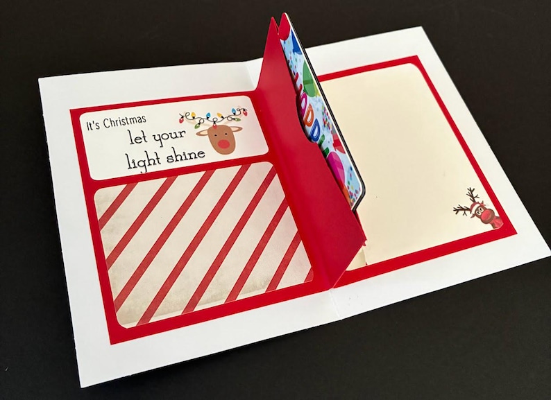 Peut inclure: Une carte de v&oelig;ux de No&euml;l avec un motif &agrave; rayures rouges et blanches et le texte "It's Christmas, let your light shine". La carte pr&eacute;sente une illustration de renne et une poche contenant une carte cadeau.