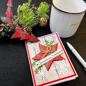 Peut inclure: Une carte de No&euml;l avec une &eacute;toile &agrave; carreaux rouges et l'inscription "Christmas Wishes" sur une banni&egrave;re blanche. La carte a un fond imitation bois et est orn&eacute;e de flocons de neige et de verdure. Une tasse blanche et un stylo sont &eacute;galement pr&eacute;sents.