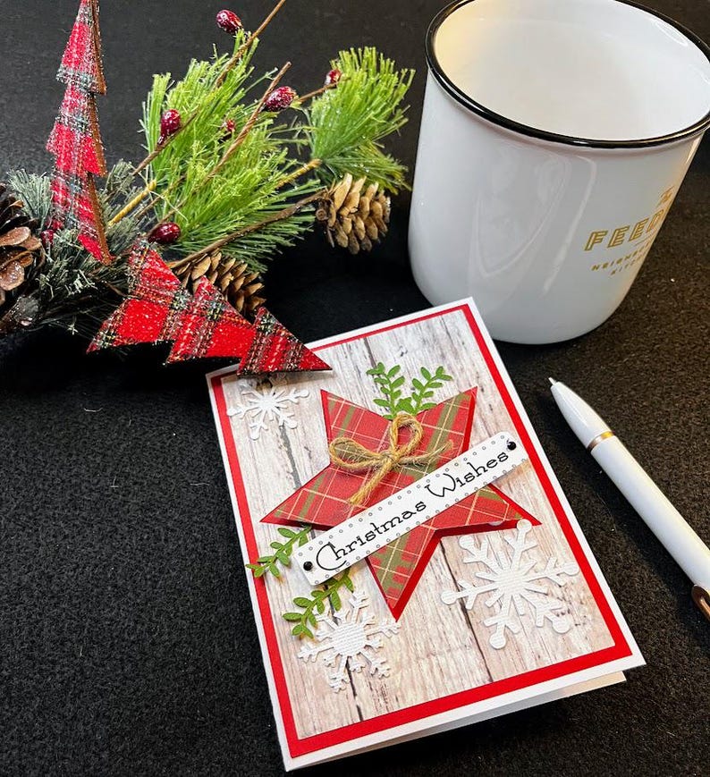 Peut inclure: Une carte de No&euml;l avec une &eacute;toile &agrave; carreaux rouges et l'inscription "Christmas Wishes" sur une banni&egrave;re blanche. La carte a un fond imitation bois et est orn&eacute;e de flocons de neige et de verdure. Une tasse blanche et un stylo sont &eacute;galement pr&eacute;sents.