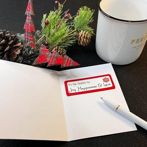 Peut inclure: Une carte de v&oelig;ux blanche ouverte avec une &eacute;tiquette rouge indiquant "It's the Season for Joy, Happiness & Love". Un stylo blanc avec des d&eacute;tails dor&eacute;s repose sur la carte. Une tasse blanche et des d&eacute;corations de No&euml;l sont en arri&egrave;re-plan.